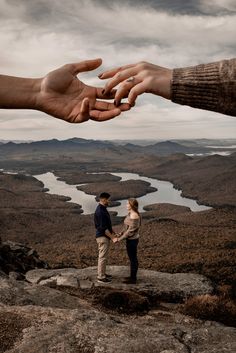 two people holding hands on top of a rocky hill with water and mountains in the background