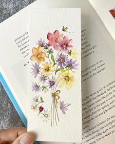 a person holding an open book with watercolor flowers on it and a ladybug in the background