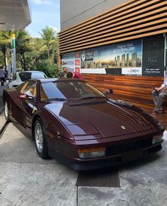 a maroon sports car parked in front of a building next to a man sitting on a bench