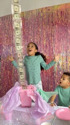 two children sitting at a table with money on top of it and balloons in the air