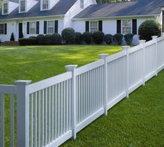 a white picket fence in front of a house with green grass and bushes around it