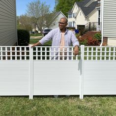 a man standing behind a white fence in front of a house with his hands on the gate