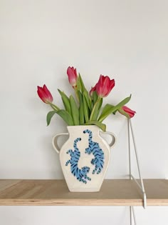 a white vase with red and blue flowers in it on top of a wooden shelf