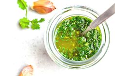 a glass jar filled with green sauce next to garlic and parsley on a white surface