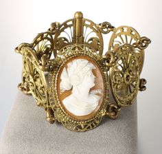 Gorgeous early 1900s link bracelet marked Made in France on the reverse. Its focal point is a right faced carved cameo of a beautiful woman that looks like a Greek or Roman Goddess.  Cameo is sitting in filigree gold tone setting and flanking it are four gold tone links with ornate open scroll work. Gold plate has aged which gives the bracelet an old kind of beauty. Length:  7 1/4 inches Width:    1 5/8 inches Clasp works very well.   Bracelet is in very good vintage condition overall and stunni 1900s Jewelry, Cameo Bracelet, Spartanburg Sc, Roman Goddess, Scroll Work, Early 1900s, Beautiful Woman, Chain Link Bracelet, Link Bracelets