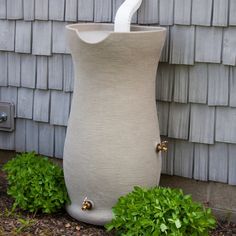 a large white vase sitting next to a green bush and a gray building with wooden shingles on it