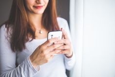 a woman looking at her cell phone while standing in front of a window and smiling