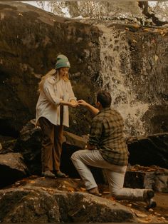 two people are standing on rocks near a waterfall and one is touching the other's hand