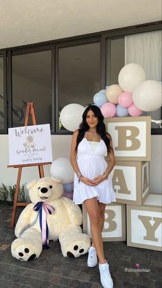 a pregnant woman standing next to a giant teddy bear in front of a baby shower sign