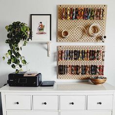 a white dresser topped with lots of drawers next to a potted plant and framed pictures