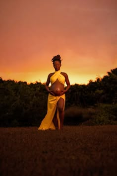 a pregnant woman in a yellow dress standing in the grass at sunset with her hands on her hips