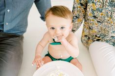 a baby sitting in front of a plate with food on it's side and people standing around him