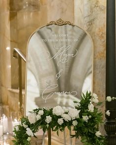 a wedding welcome sign with white flowers and greenery in front of a large mirror