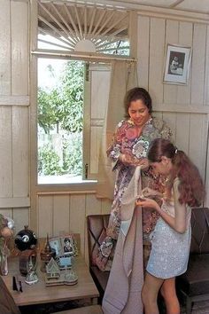 two women standing in a living room next to a table and chair with flowers on it