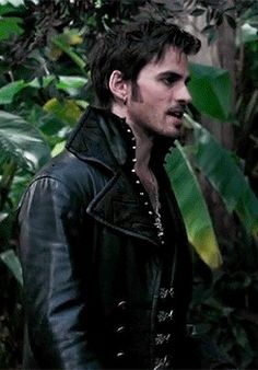 a man standing in front of green plants and trees wearing a leather jacket with his hands on his hips