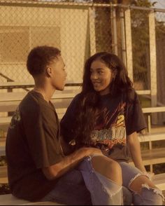 two people sitting on a bench talking to each other in front of a chain link fence