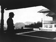 a black and white photo of a man standing in front of a building with mountains behind him