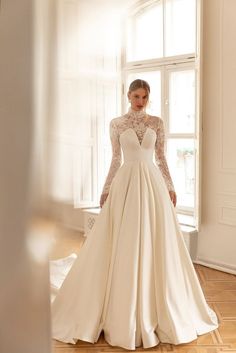 a woman in a wedding dress standing by a window with her hands on her hips