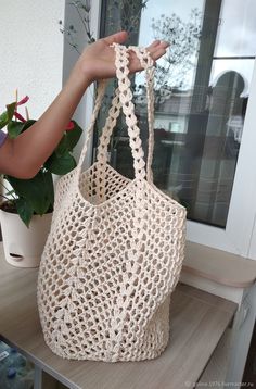 a hand holding a crocheted bag on top of a shelf next to a potted plant