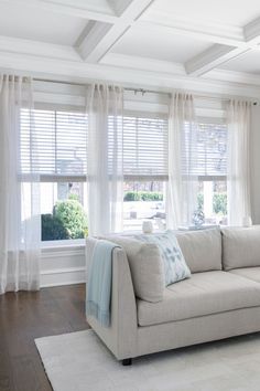 a living room filled with furniture and windows covered in white drapes on top of wooden flooring