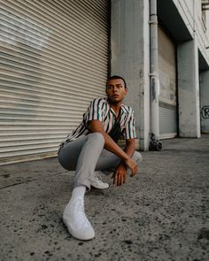 a man sitting on the ground in front of a garage door wearing white sneakers and a striped shirt