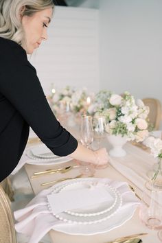 a woman is setting the table for a formal dinner with white flowers and greenery