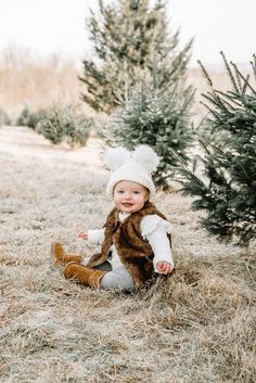 a baby sitting on the ground in front of some christmas trees wearing a hat and vest