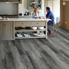 a man and woman sitting at a counter in a kitchen with grey wood flooring