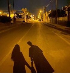 two people walking down the street at night with their shadows on the road and buildings in the background