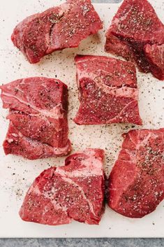 raw steaks on a cutting board with seasoning sprinkled over them and sitting on a white surface