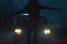 a woman standing in the street at night with her arms spread out and two cars behind her