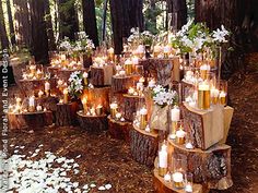 many candles are lit in the woods with white flowers and greenery on each tree stump