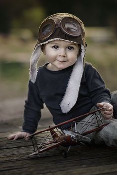 a small child wearing a hat and goggles sitting on the ground with a toy airplane