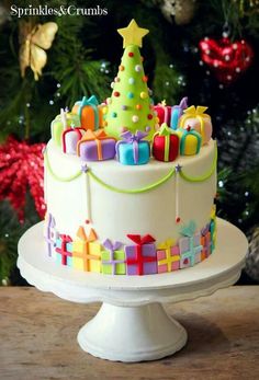 a brightly decorated christmas cake on a table in front of a christmas tree with presents