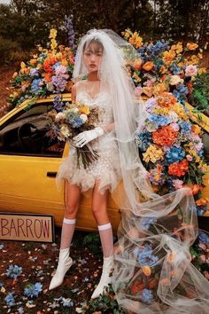 a woman standing in front of a yellow car with flowers on the hood and veil