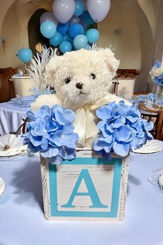 a teddy bear is sitting in a box with blue flowers on the table at a baby's first birthday party