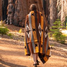 a woman walking down a dirt road with a blanket on her back and trees in the background