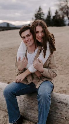 a young man and woman sitting on a log together, smiling at the camera with their arms around each other