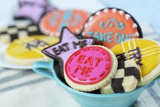 some decorated cookies in a blue bowl on a striped table cloth with the words eat me