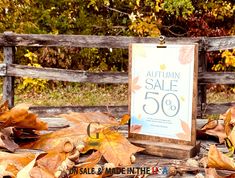 an autumn sale sign sitting on top of leaves in front of a wooden fence with fall foliage around it