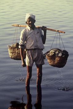 a man standing in the water with two baskets on his shoulders and one holding a stick