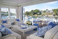 a living room filled with white furniture next to a large window covered in blue and white pillows