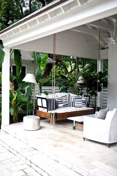 a porch swing with cushions on it in front of a white house and some trees