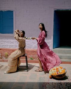 two women in long dresses are sitting on a chair and one woman is holding flowers