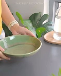 a woman holding a green bowl on top of a table next to a plant and a glass bottle