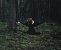 a woman with red hair sitting on the ground next to a tree in a forest