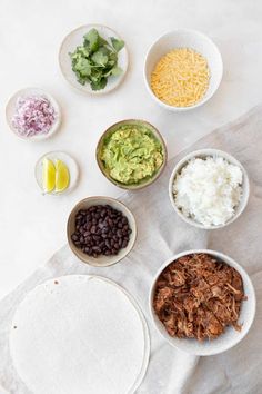 bowls filled with different types of food sitting on top of a white table cloth next to tortilla shells