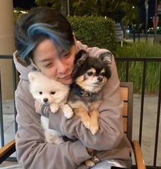 a woman sitting on a bench holding two small white and brown dogs in her arms