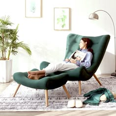 a woman sitting in a green chair reading a book on the floor next to a potted plant