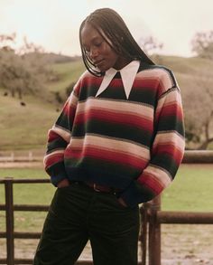 A person stands outdoors near a wooden fence in front of a hilly landscape with scattered trees. They are wearing The Bubble Pullover by The Great Inc., featuring painterly stripes and a white collar, paired with dark pants. Their head is slightly tilted down, and their long hair is styled in braids. Scented Body Oils, Voluminous Sleeves, Denim Hat, The Bubble, Kids Outerwear, Baby Alpaca, Soft Wool, Knit Shirt, Wool Yarn